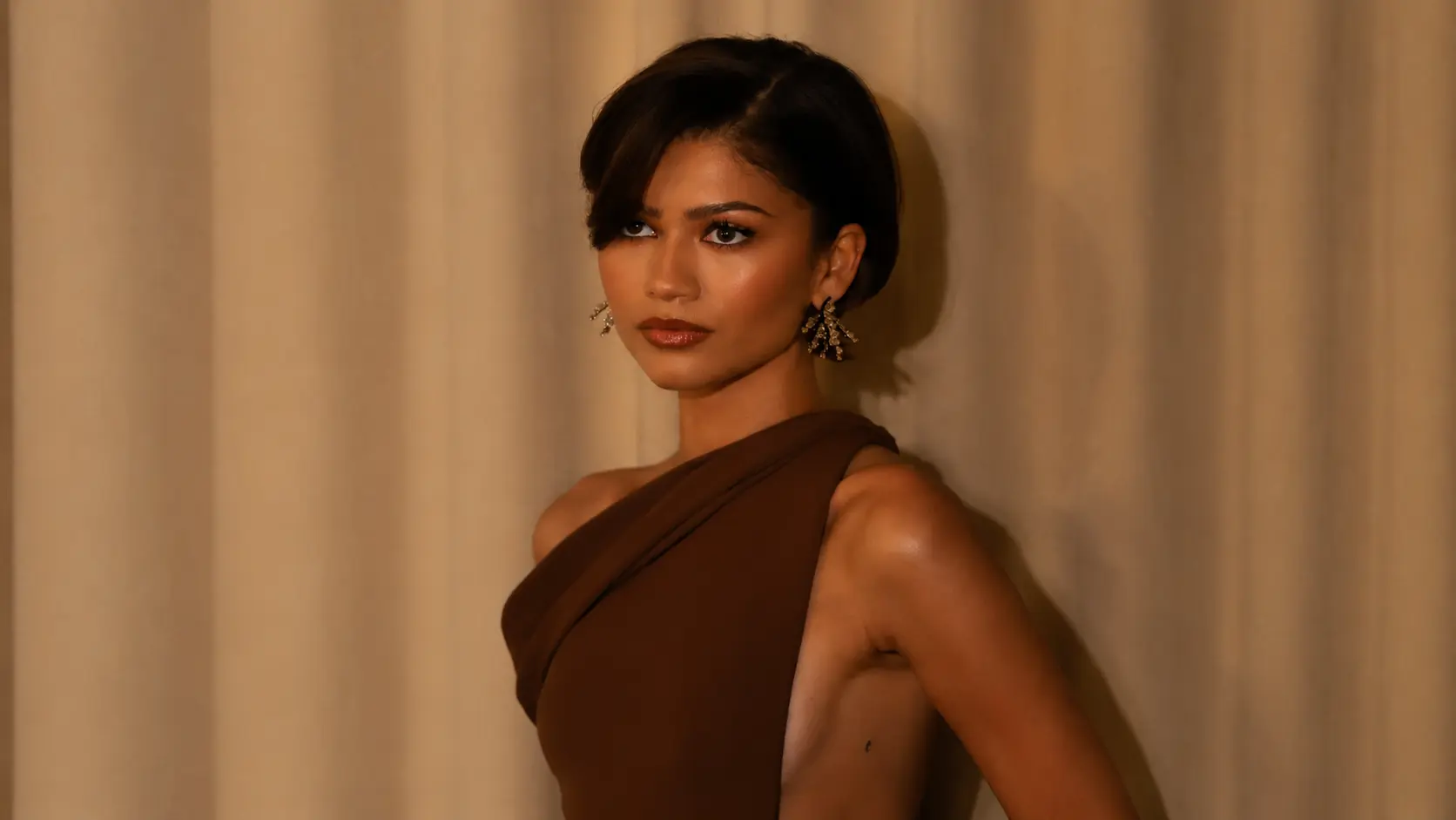 zendaya with short dark hair wearing a brown one-shoulder dress and earrings, posing in warm lighting against a beige background.