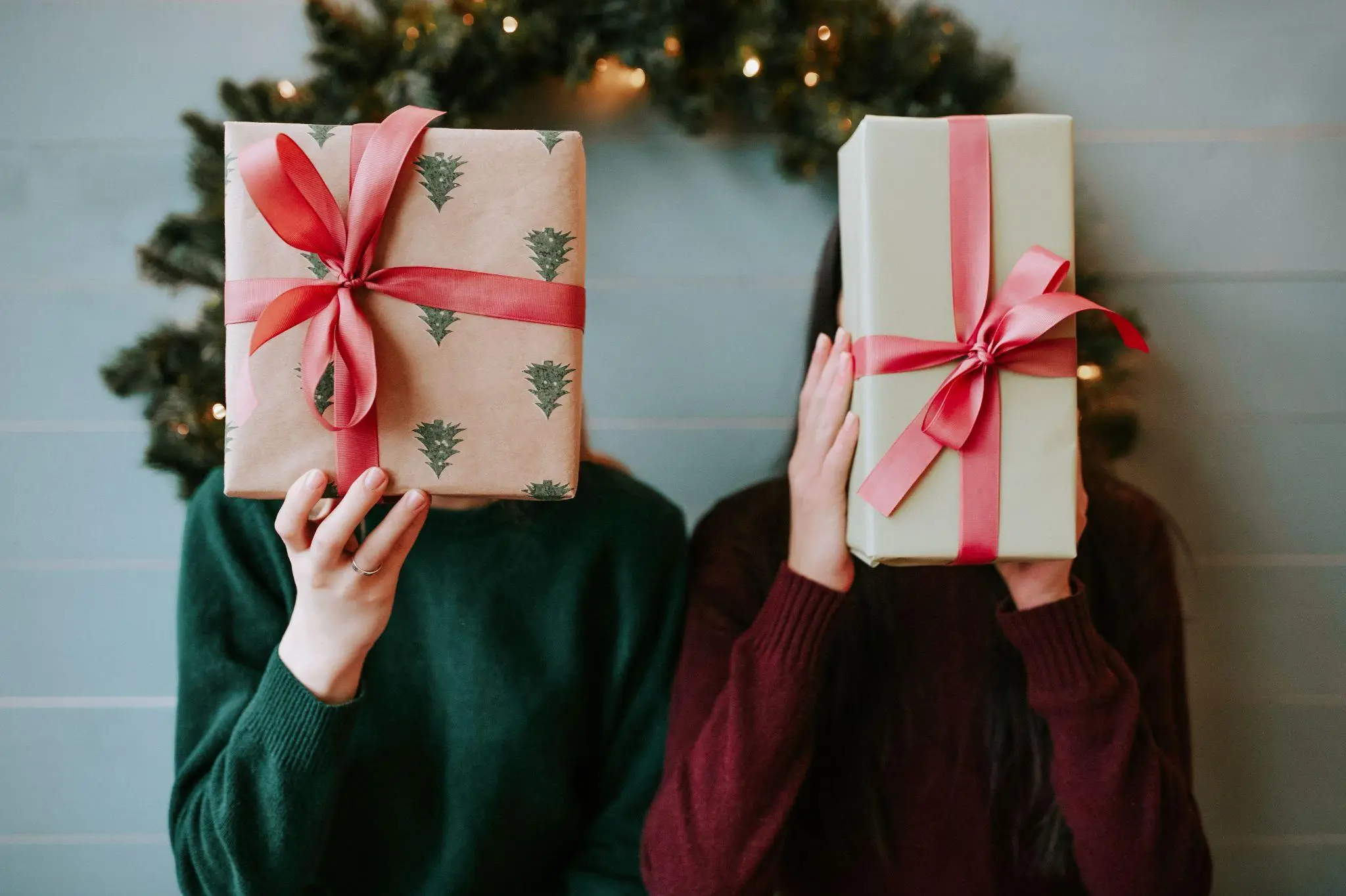 Two people holding wrapped gifts with red bows indoors against holiday decor backdrop