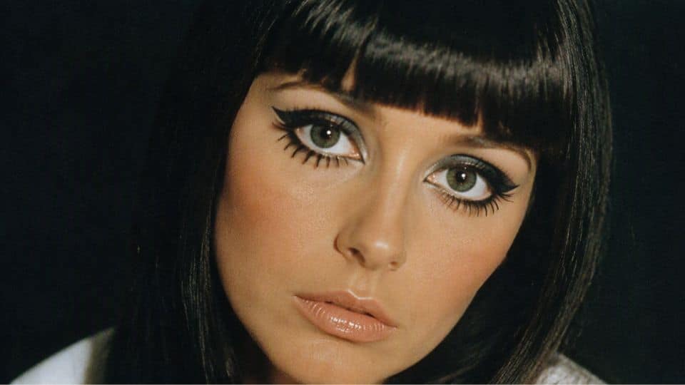 Cher inspired 60s makeup with long graphic eyeliner, dramatic lashes, bare lids, and glossy nude lips