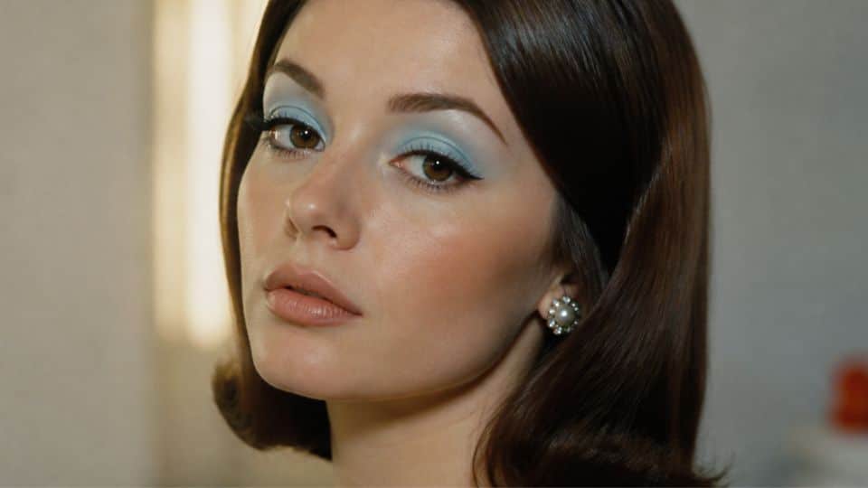 1960s makeup look with pastel blue eyeshadow, black liner, voluminous lashes, and soft nude lips