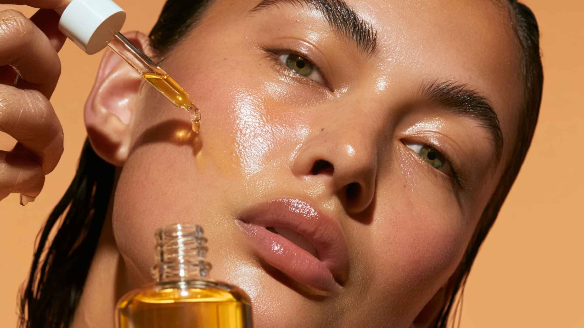 A close-up beauty skincare scene featuring a person applying golden face oil with a glass dropper onto glowing, hydrated skin.