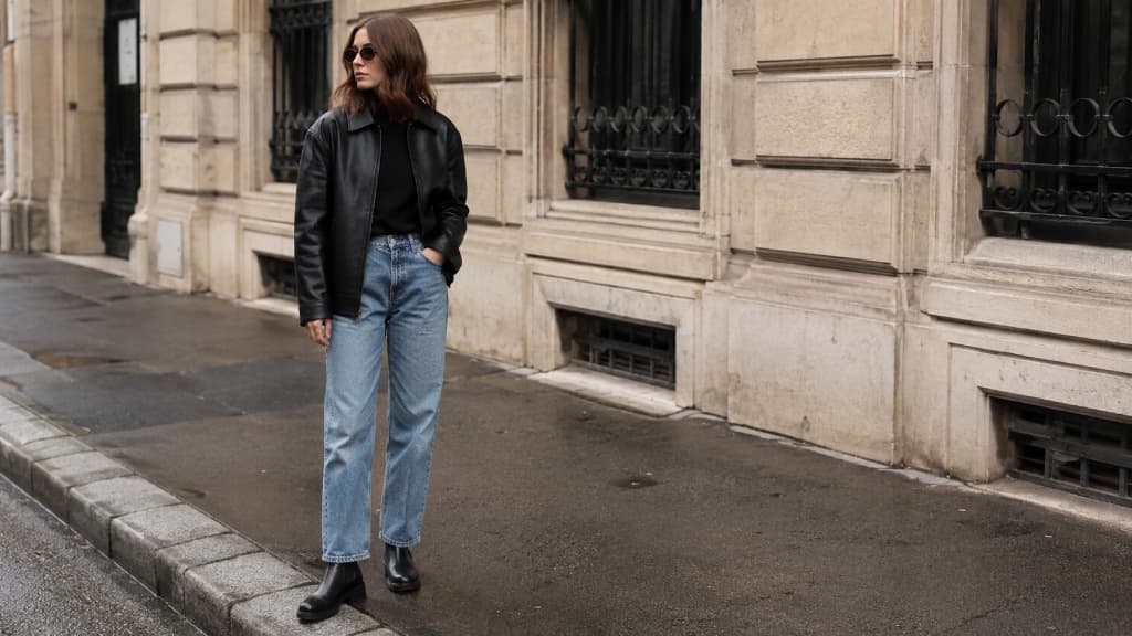A woman in sunglasses, a black leather jacket, blue jeans, and black boots standing on a city street.