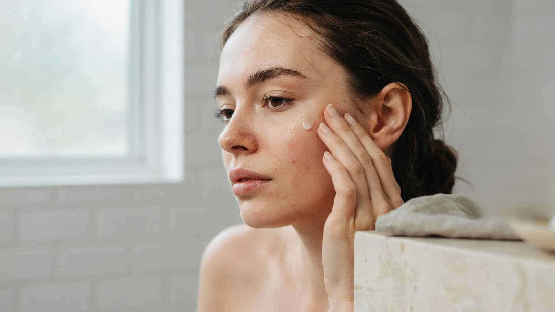 Woman checking her skin in the mirror while holding a foundation bottle, illustrating whether makeup causes acne.