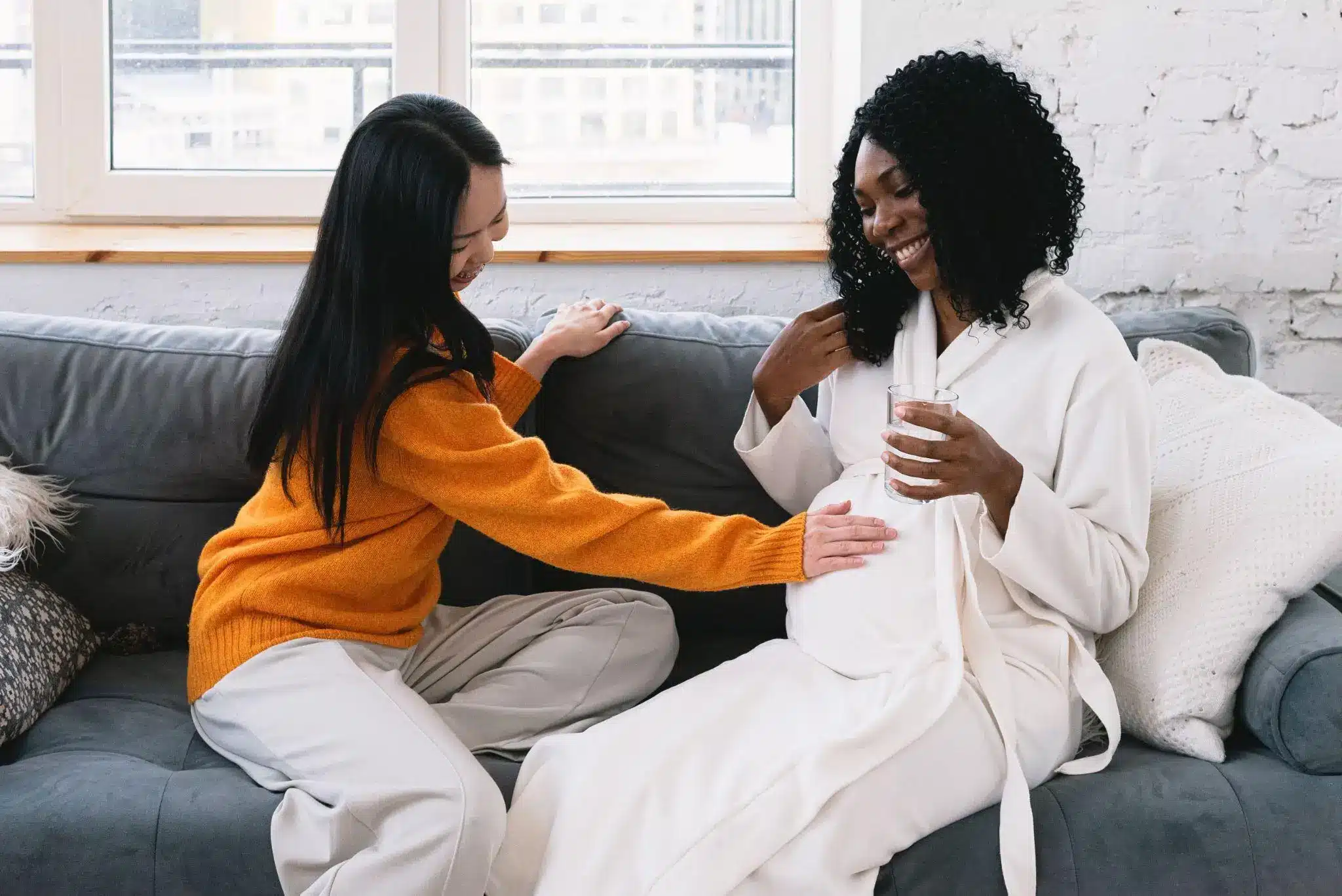 Two women sitting on a couch, one touching the other's pregnant belly, in a bright room