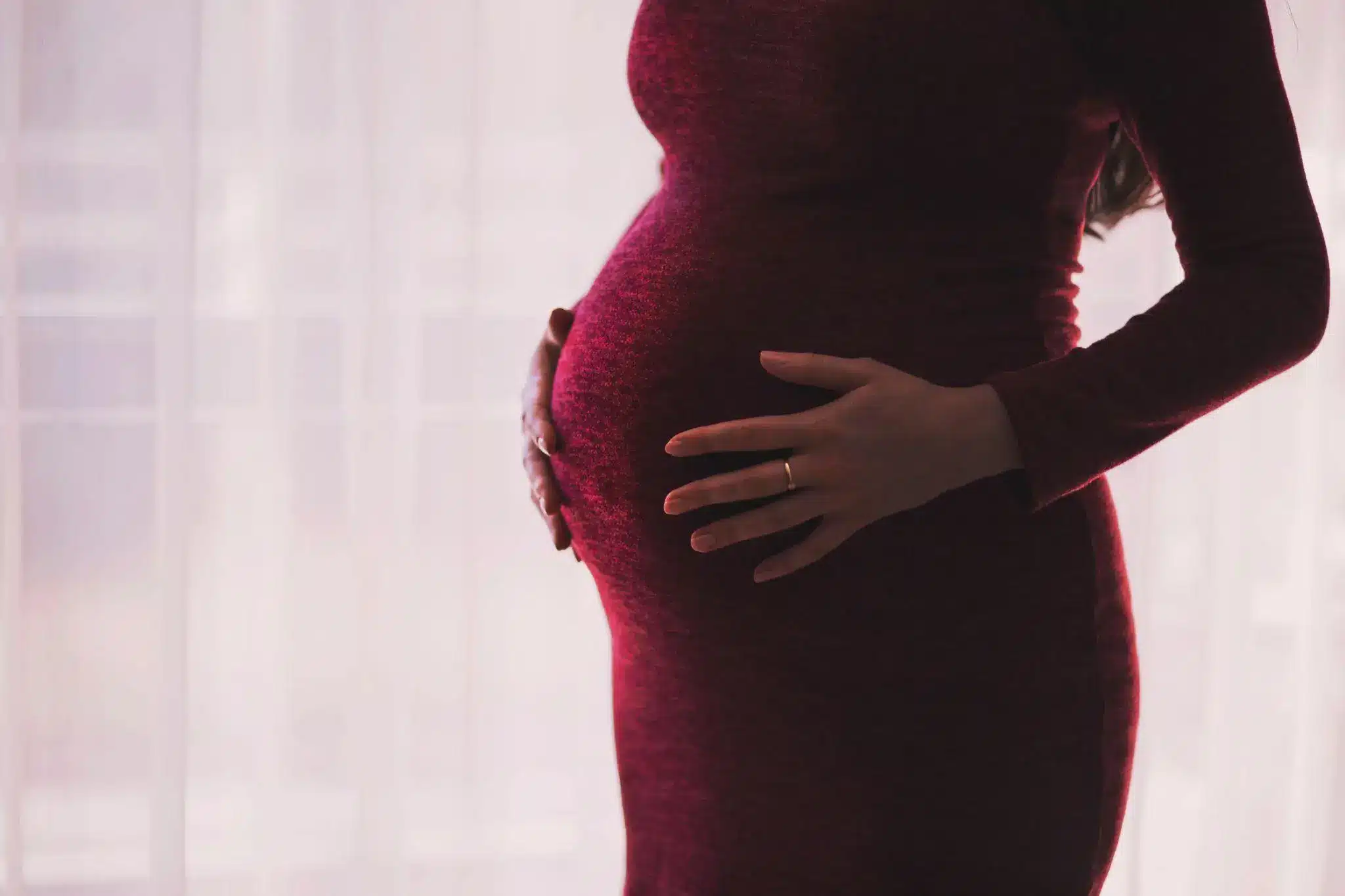 Pregnant woman in burgundy dress gently holding her belly in soft lighting