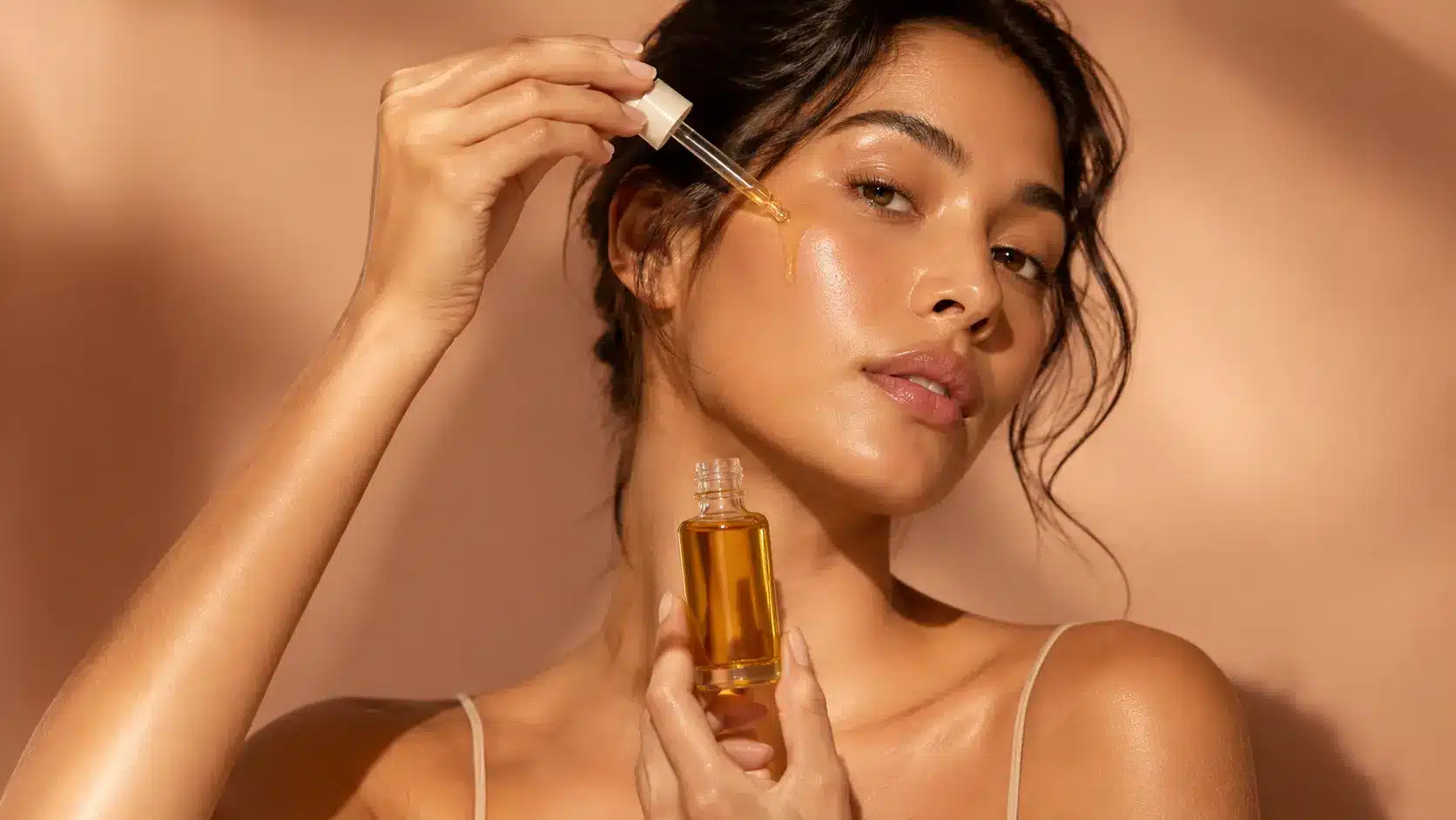 Close up beauty portrait of a woman applying facial oil with a dropper beside an unbranded amber glass skincare bottle