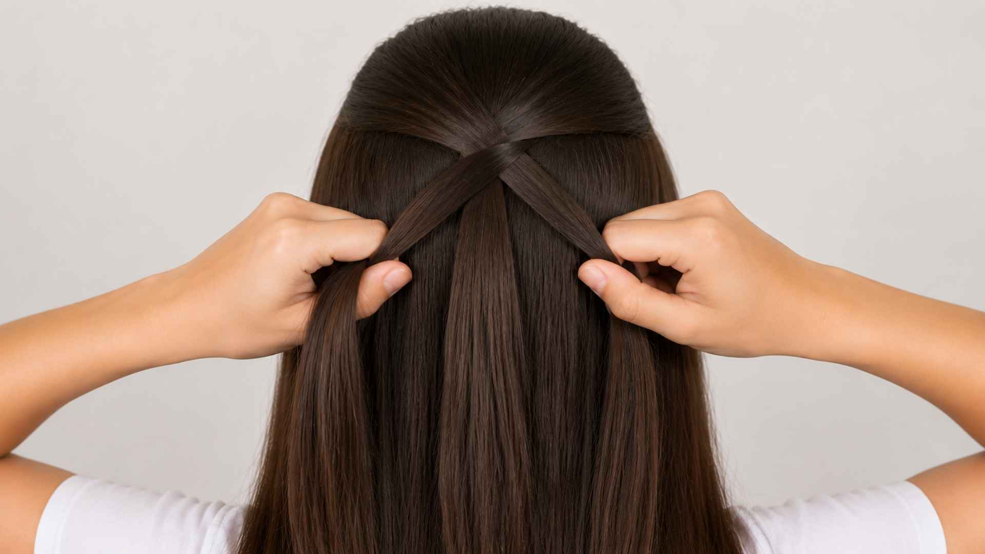 Hands crossing three hair strands together at the back while forming the base of a French braid pattern