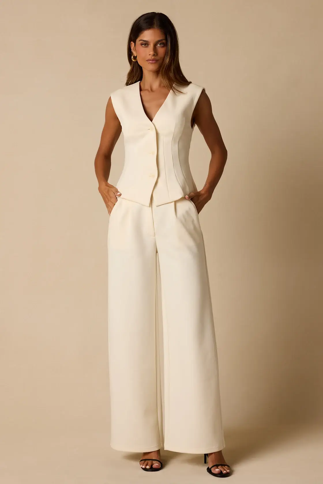 Woman in cream-colored tailored vest and wide-leg pants standing against beige background