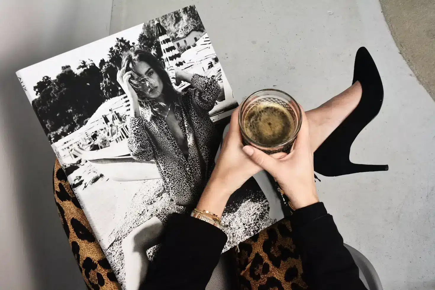 Person holding coffee above fashion magazine wearing black high heels and leopard print pants