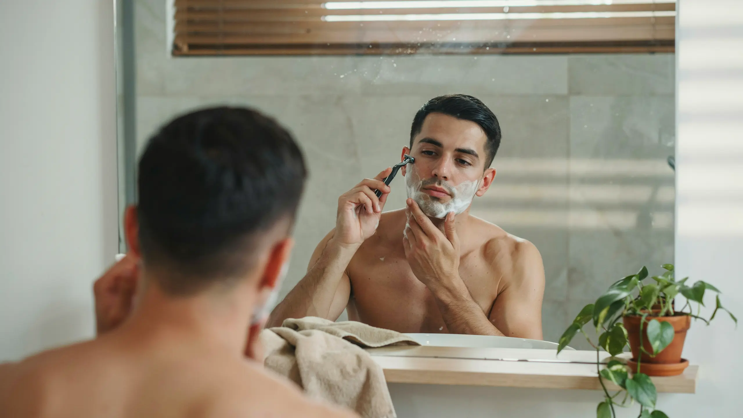Man applying shaving cream and grooming his face with a razor in front of a bathroom mirror