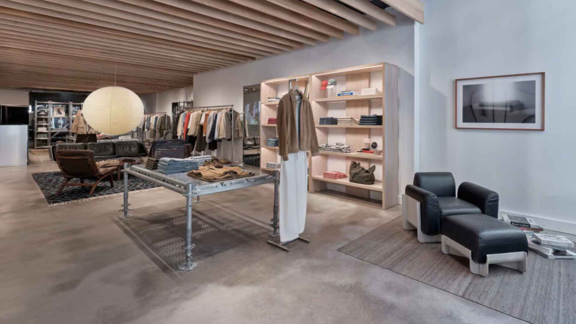 Modern fashion store interior with clothing racks, wooden shelving, display tables, and black lounge chairs.