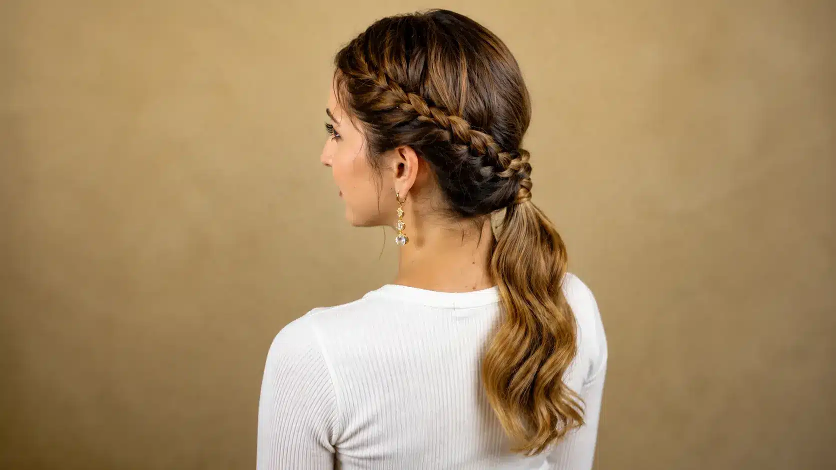 Woman from behind featuring a side Dutch braid pulled back into a wavy low ponytail against a neutral background.