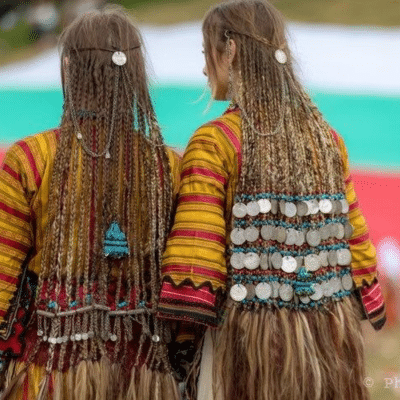 Woman wearing a traditional braided hairstyle with a long braid, patterned shawl, and fur garment outdoors.