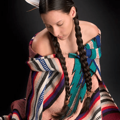 Woman wearing two long braided hairstyles with a feather accessory and traditional patterned clothing.