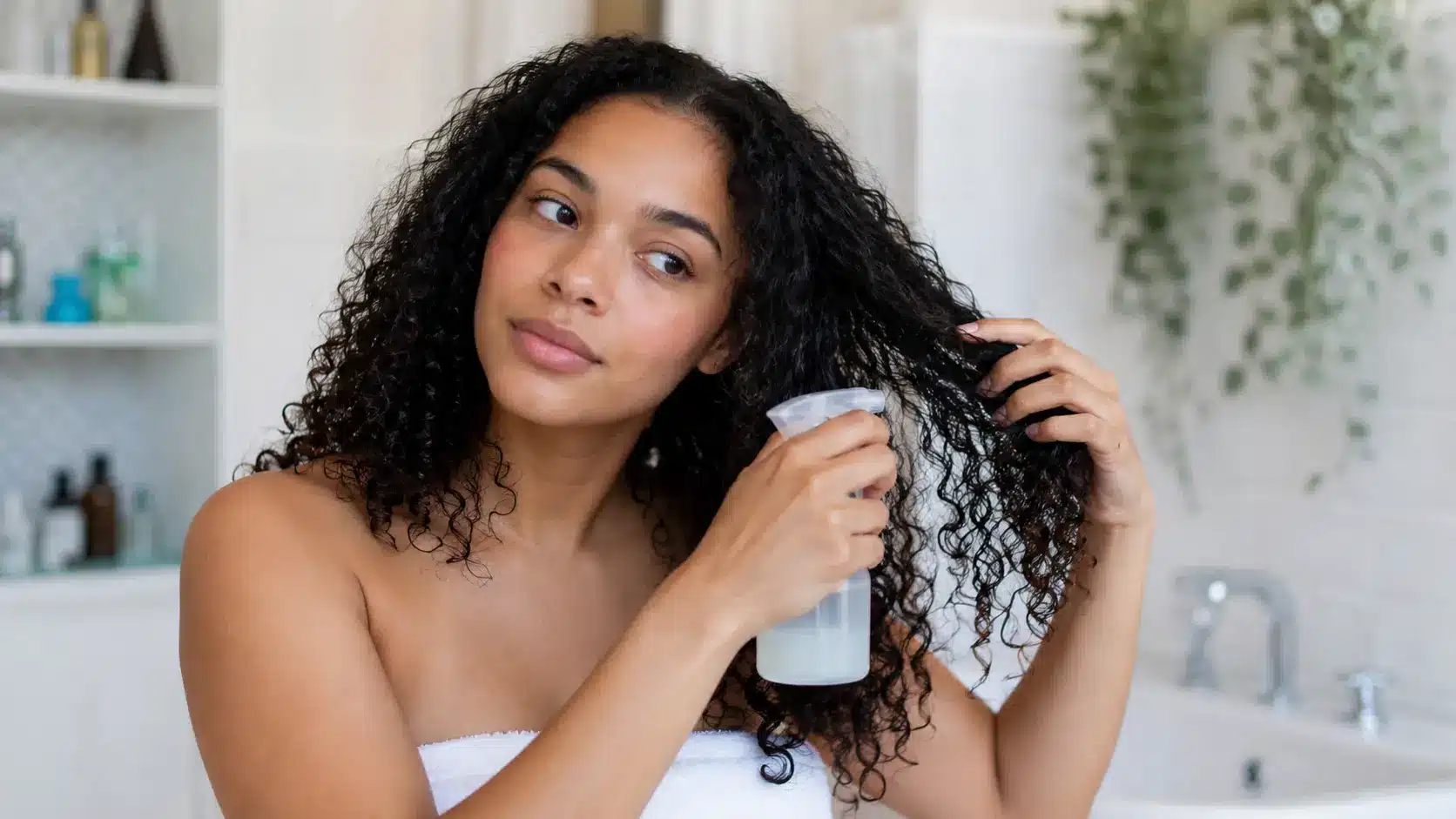 Young woman with curly hair sprays rice water through damp curls during a simple hair care routine in a bright bathroom