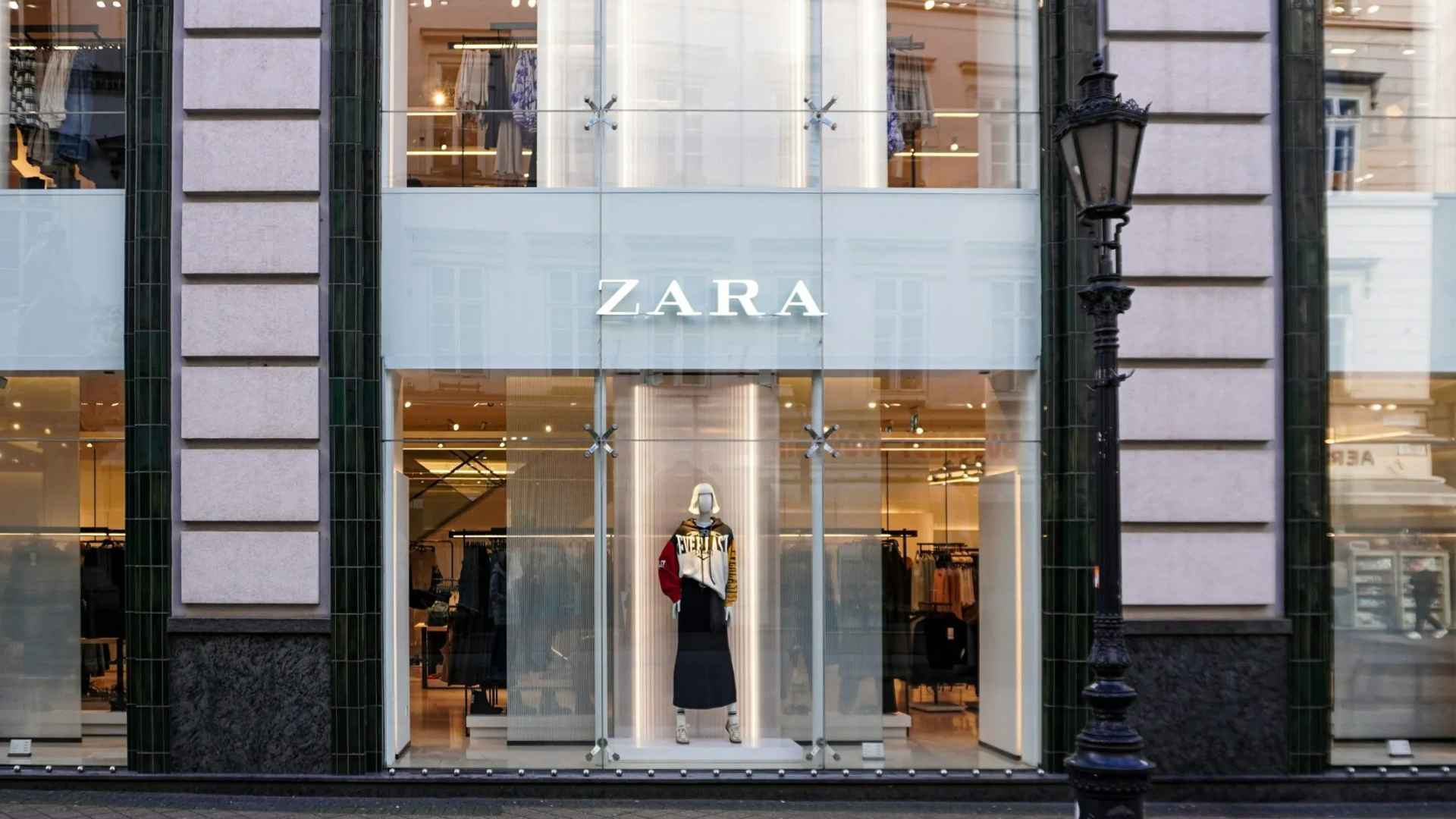 Zara storefront with a glass entrance, illuminated displays, clothing racks, and a mannequin visible inside the fashion store.