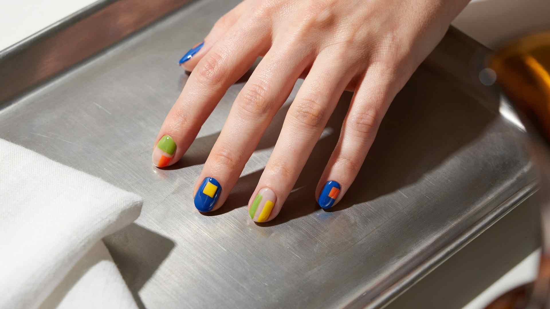 Cobalt blue geometric nails mix lime orange and yellow shapes across short tapered square tips with glossy polish