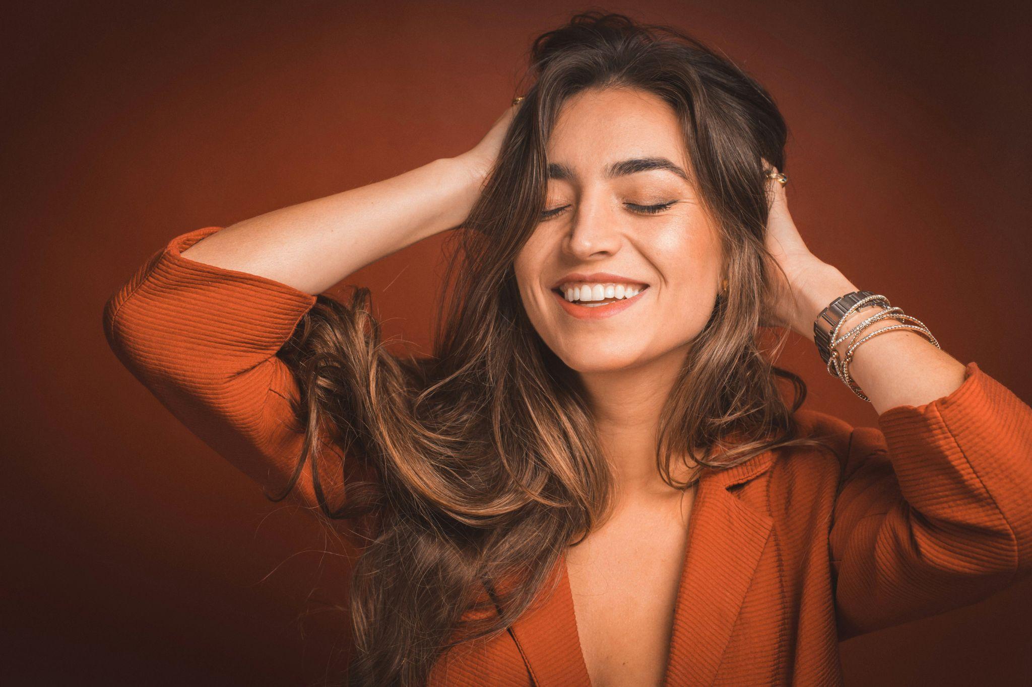 Woman smiling with eyes closed, wearing orange blazer, hands in hair against terracotta background