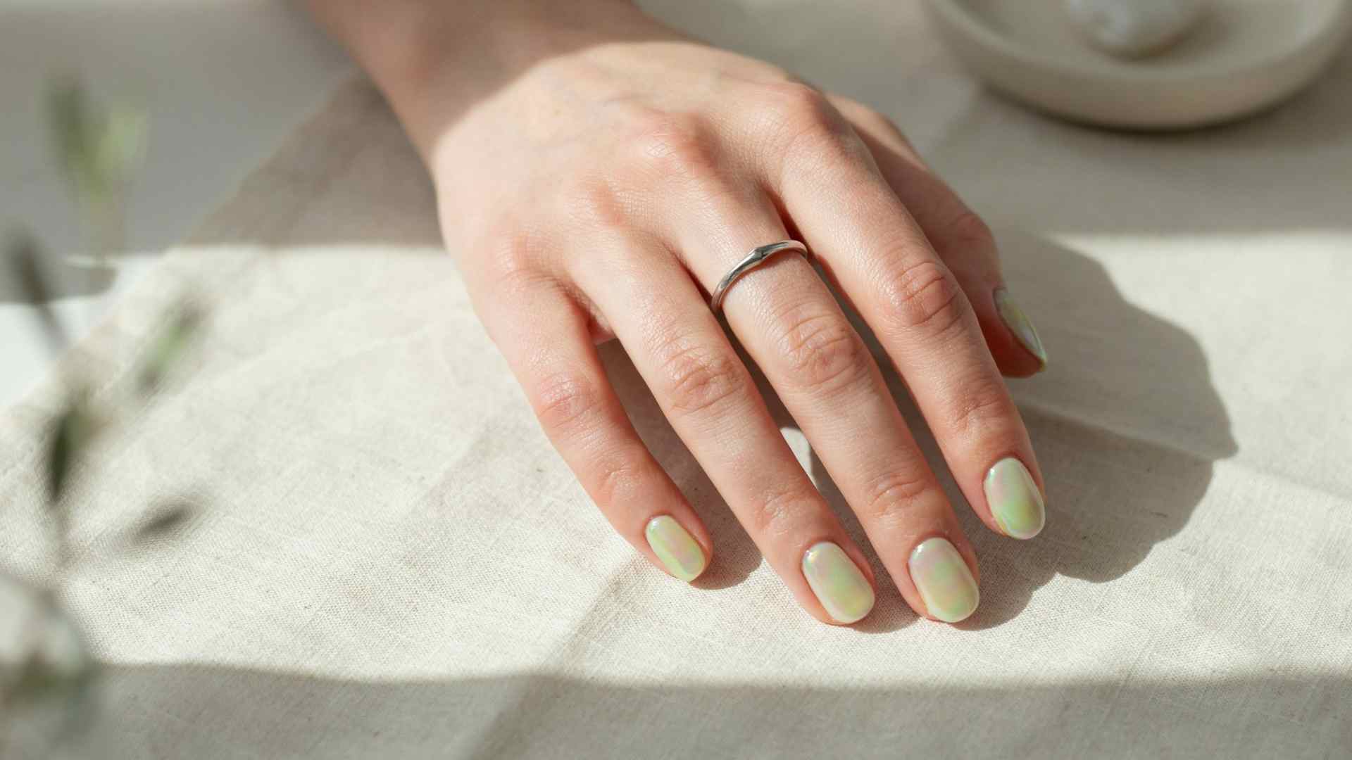 Lime green chrome almond nails with a pearly reflective finish photographed in soft daylight against pale cream linen