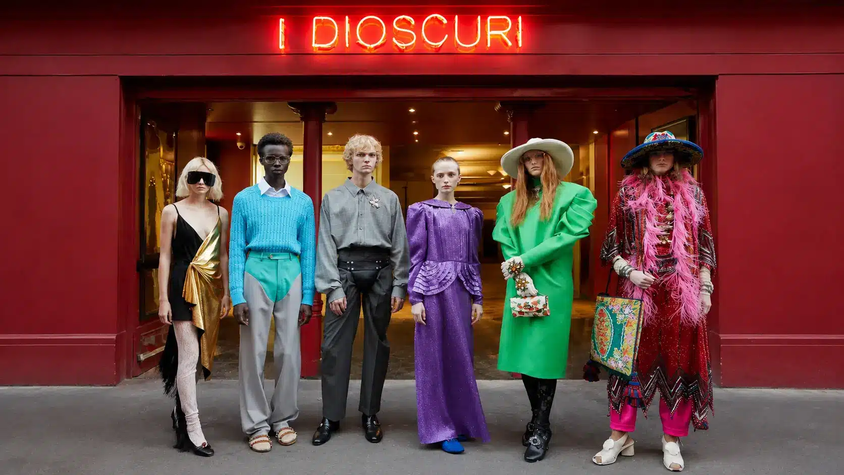 Fashion models in colorful couture outfits standing outside a red storefront with neon sign and dramatic editorial styling.