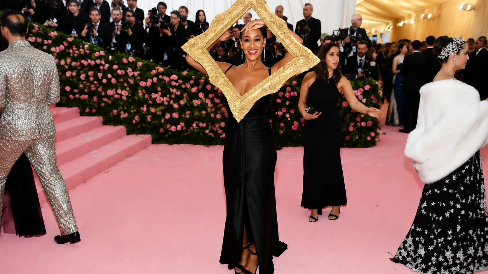 Camp fashion moment with Tracee Ellis Ross wearing a black gown and oversized gold frame on a glamorous pink carpet
