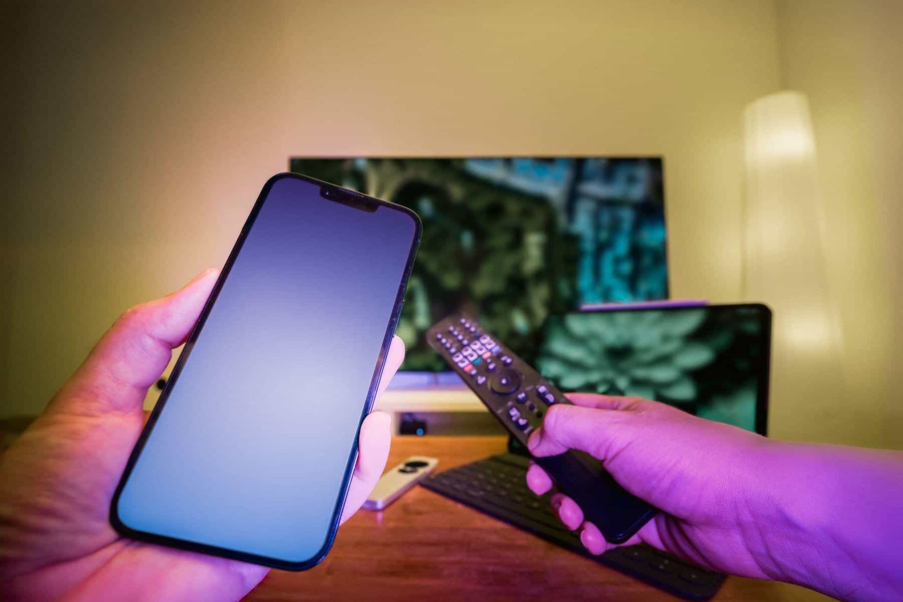 Hands holding smartphone and remote control in dimly lit room with TV and tablet in background