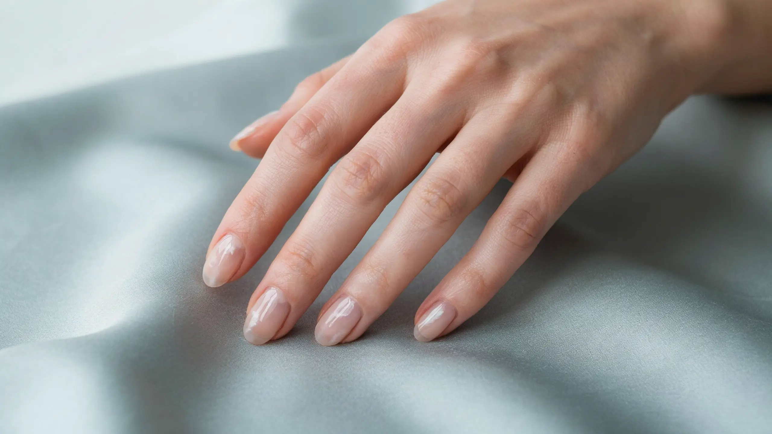 Hand with glossy cloud inspired nude nails resting on smooth gray satin, with soft cool light and gentle flowing shadows