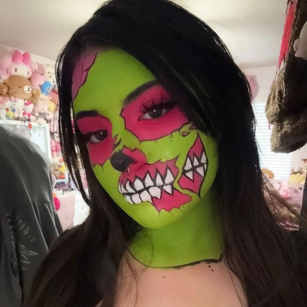 Cute zombie makeup with a grey green face, shadowed eyes, light scratches, a small blood smear, and muted grey lips