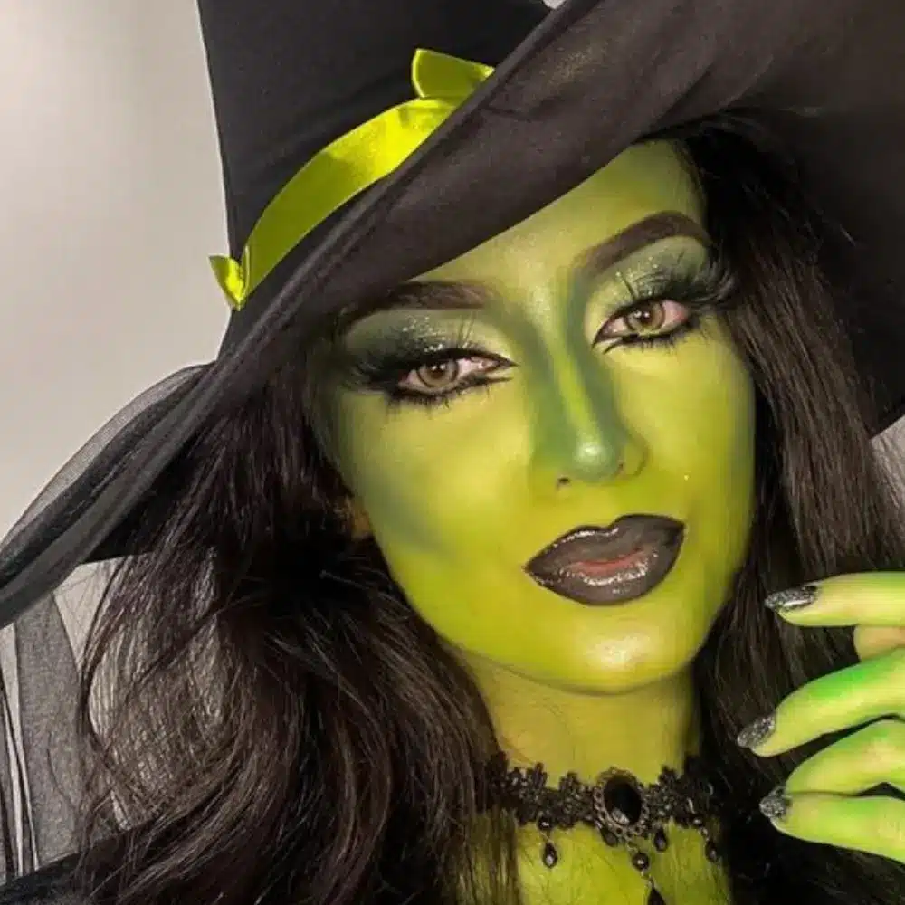 Elphaba makeup with vivid green skin, sculpted cheeks, dark brows, winged liner, and a softly defined warm nude lip