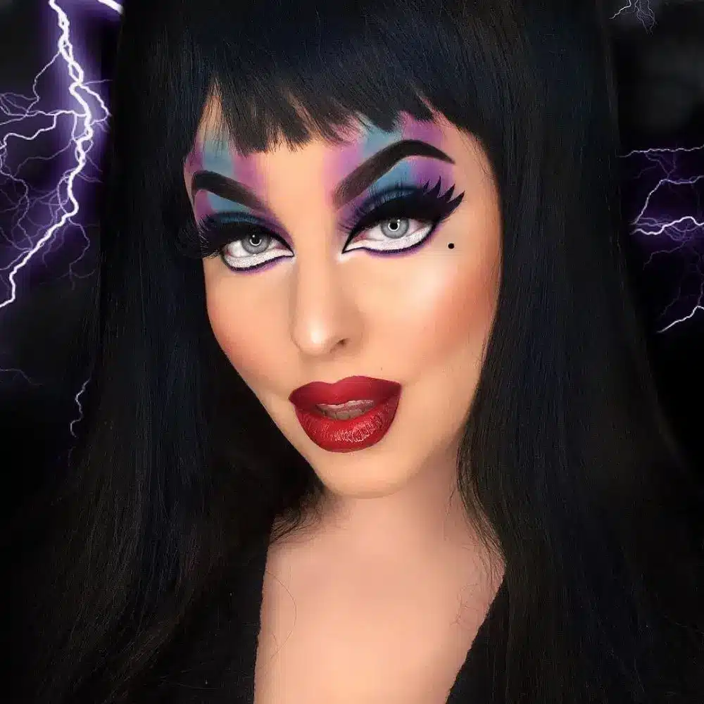 Elvira makeup with a pale matte base, contoured cheeks, arched dark brows, long black wings, and deep wine red lips