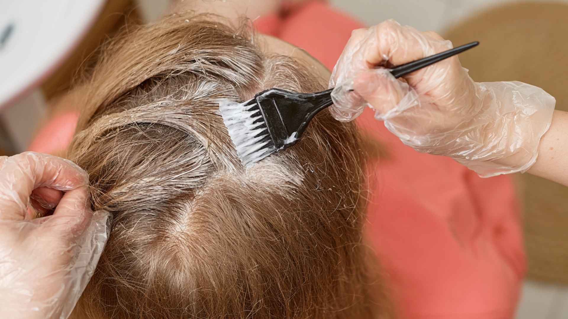 Hair colorist applying dye to a client's roots with a brush while wearing protective gloves.
