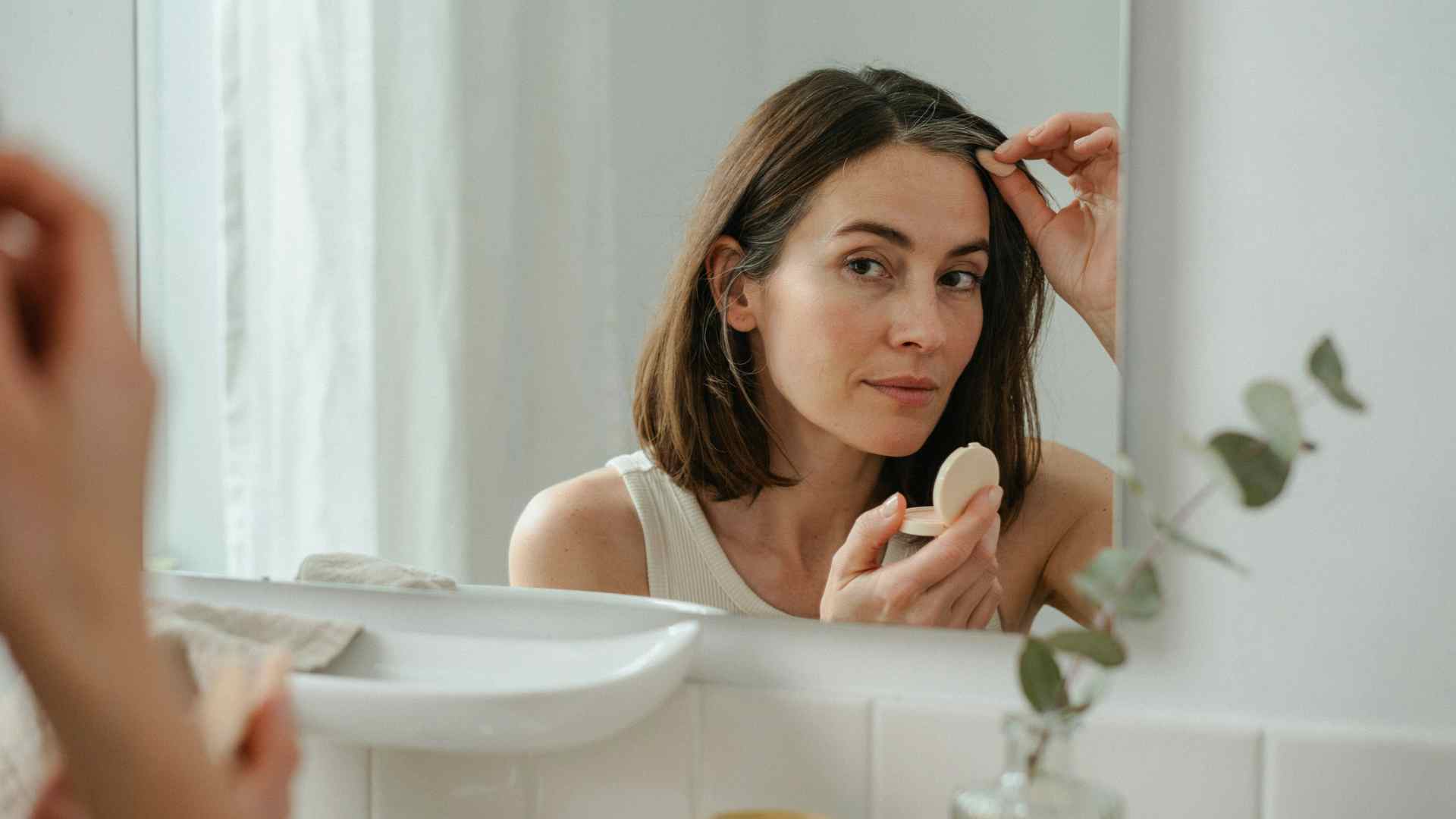 Woman applying root cover up powder near her hair part while checking gray roots in a mirror with natural bathroom lighting