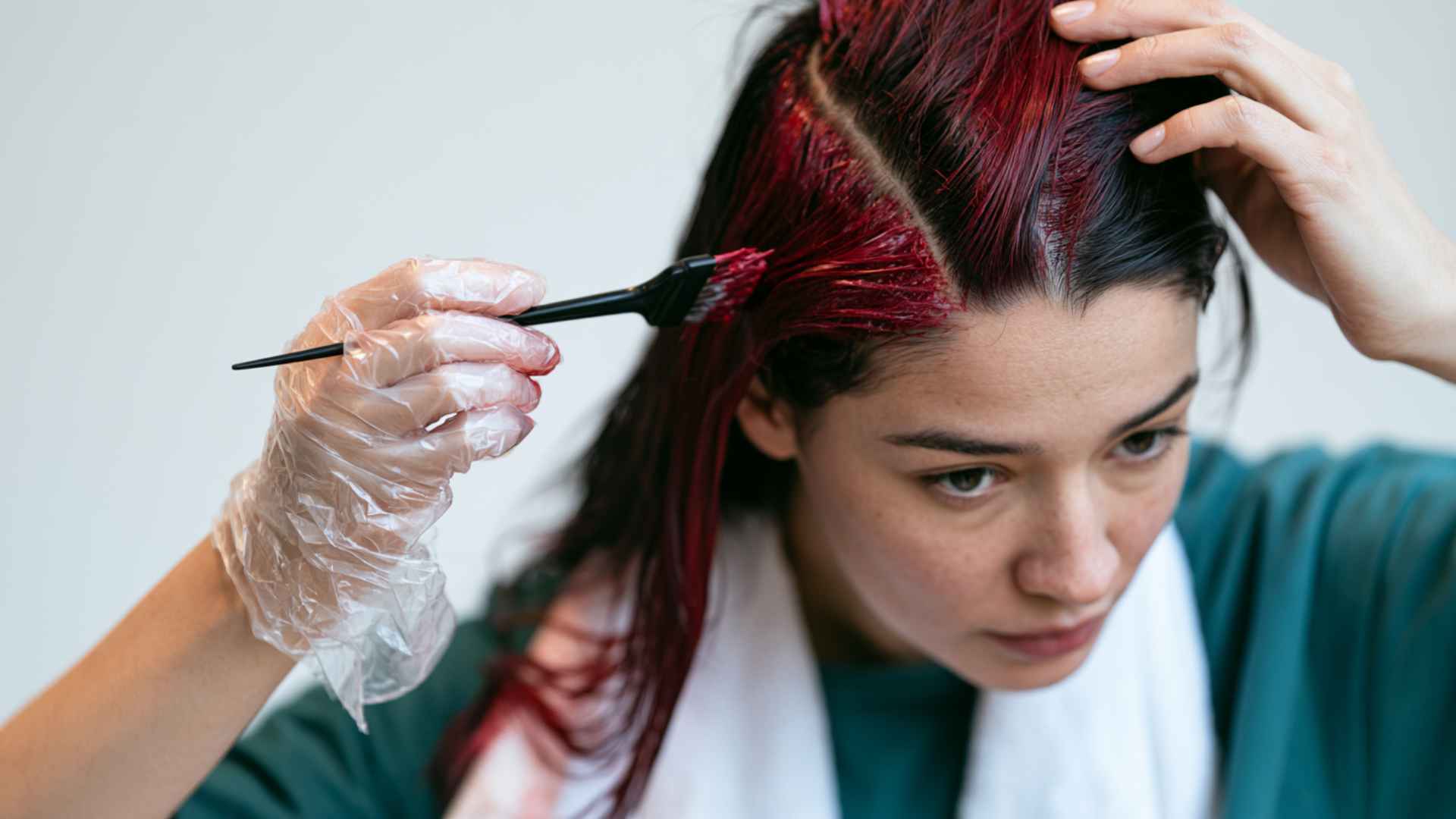 Person applying red box dye to hair roots while wearing protective gloves during at home hair coloring.