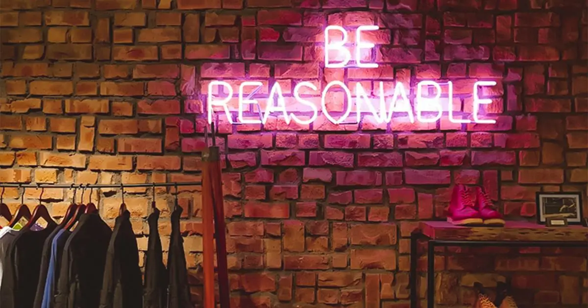 Neon sign saying "Be Reasonable" on brick wall above clothing rack and shoes