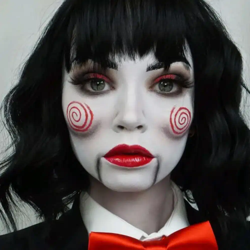 Jigsaw makeup with grey lifeless skin, dark shadowed eyes and temples, muted dark lips, and a thin fake blood trail