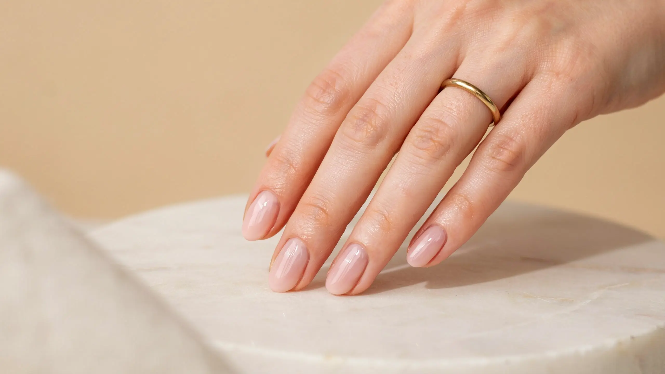 Hand with milky pink polished nails and a slim gold ring resting on a white stone surface, in warm natural sunlight glow