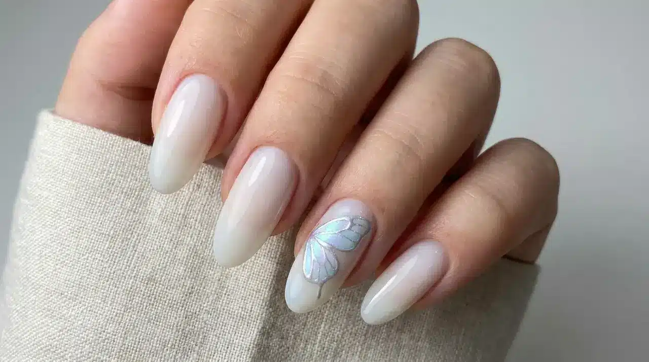 Long almond shaped milky white nails with a delicate iridescent butterfly accent design on one nail over fabric background