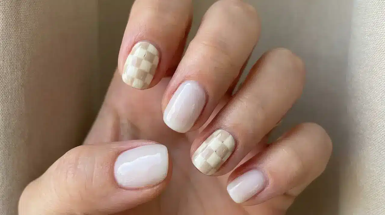 Short square milky white nails with subtle checkerboard accent designs on a hand in a minimalist manicure style