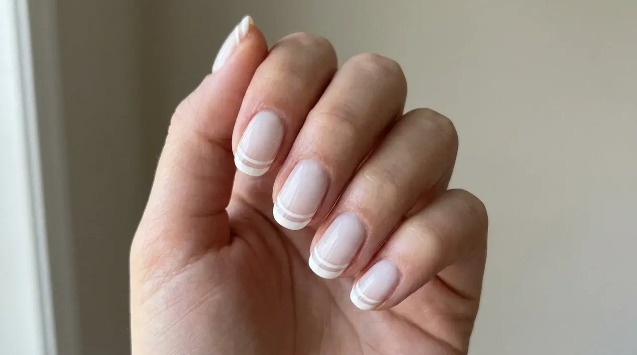 Short square milky white nails with delicate double French tip accents on a hand in a minimalist manicure style