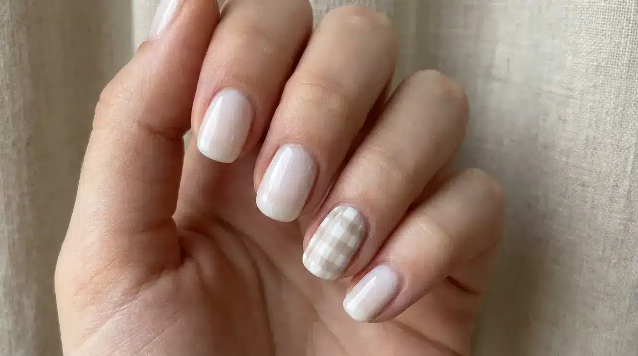 Short square milky white nails with a subtle milky gingham picnic accent design on one nail over a textured fabric background