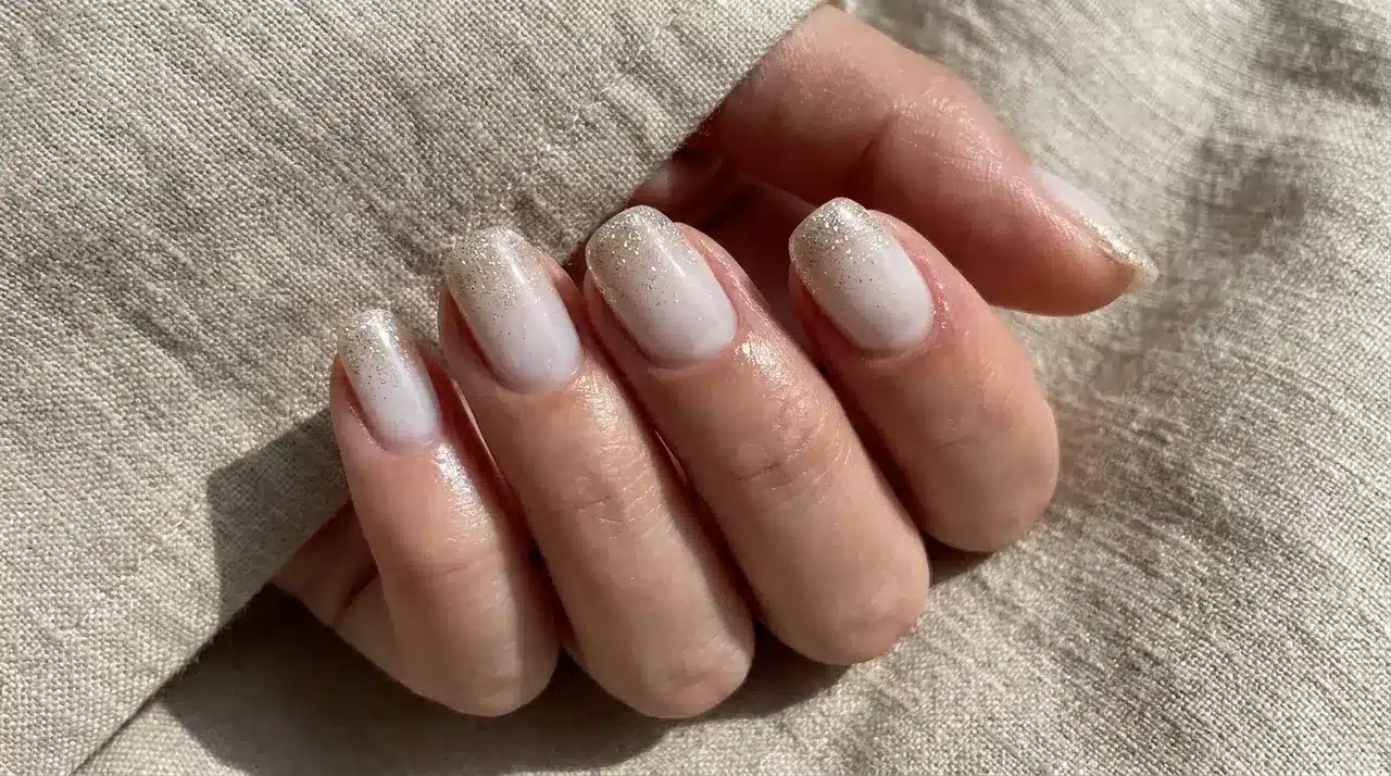 Short square milky white nails with subtle silver glitter tips on a hand resting over textured fabric background