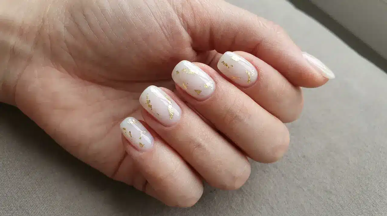 Short oval milky white nails with delicate gold foil accents and glossy finish on a hand resting over a neutral surface