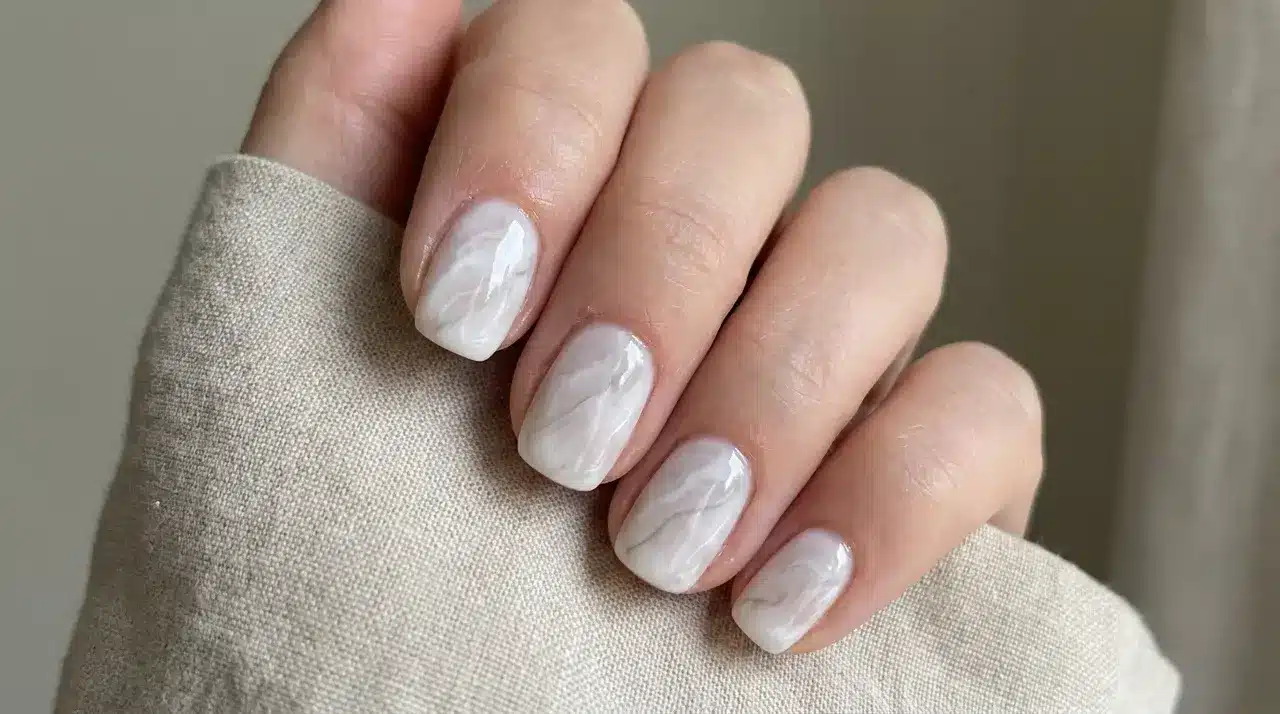 Short square milky white nails featuring a subtle marble swirl design with a glossy finish on a hand over textured fabric background