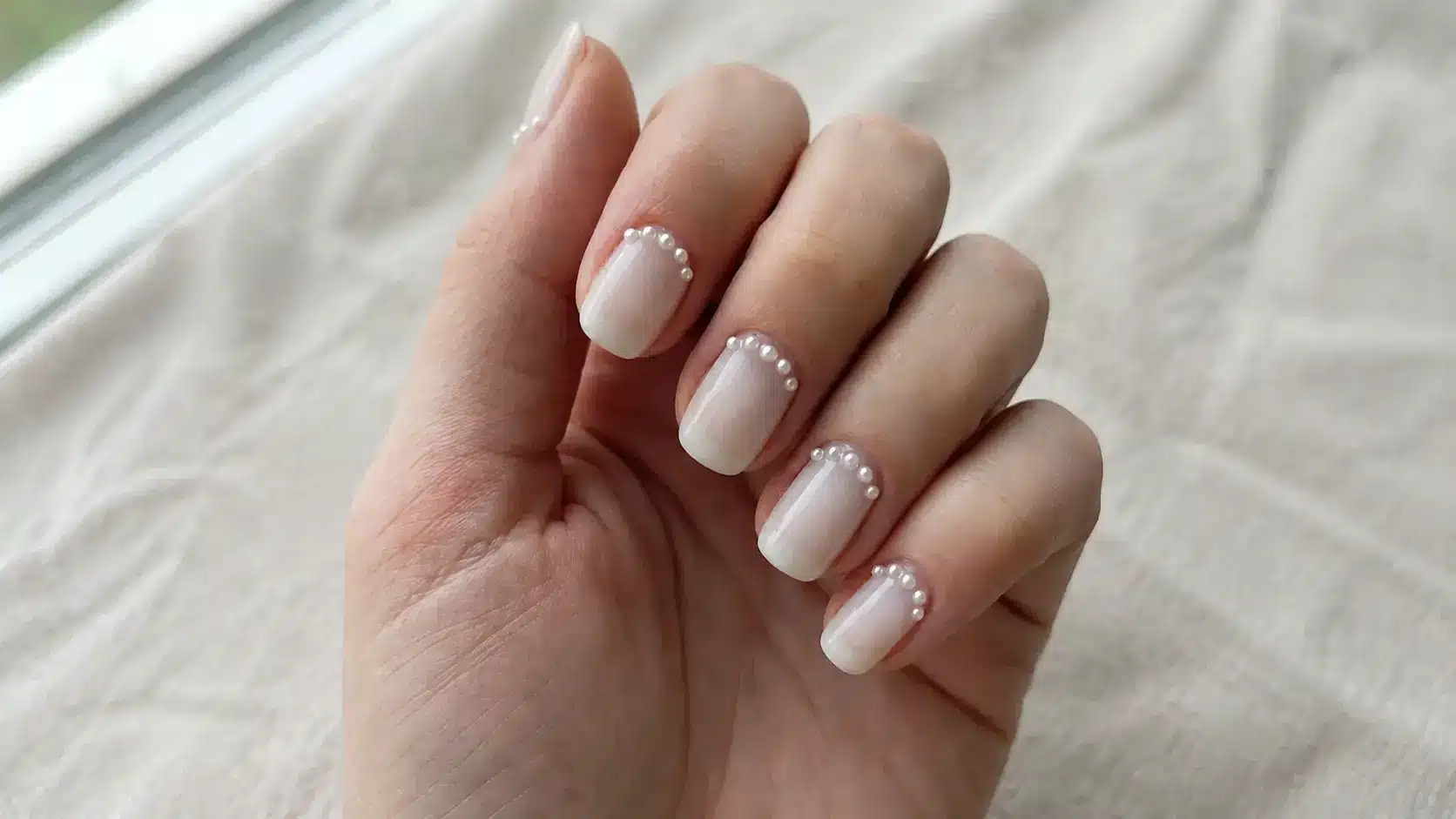 Short square milky white nails with pearl accents along the cuticles on a hand resting against a soft fabric background