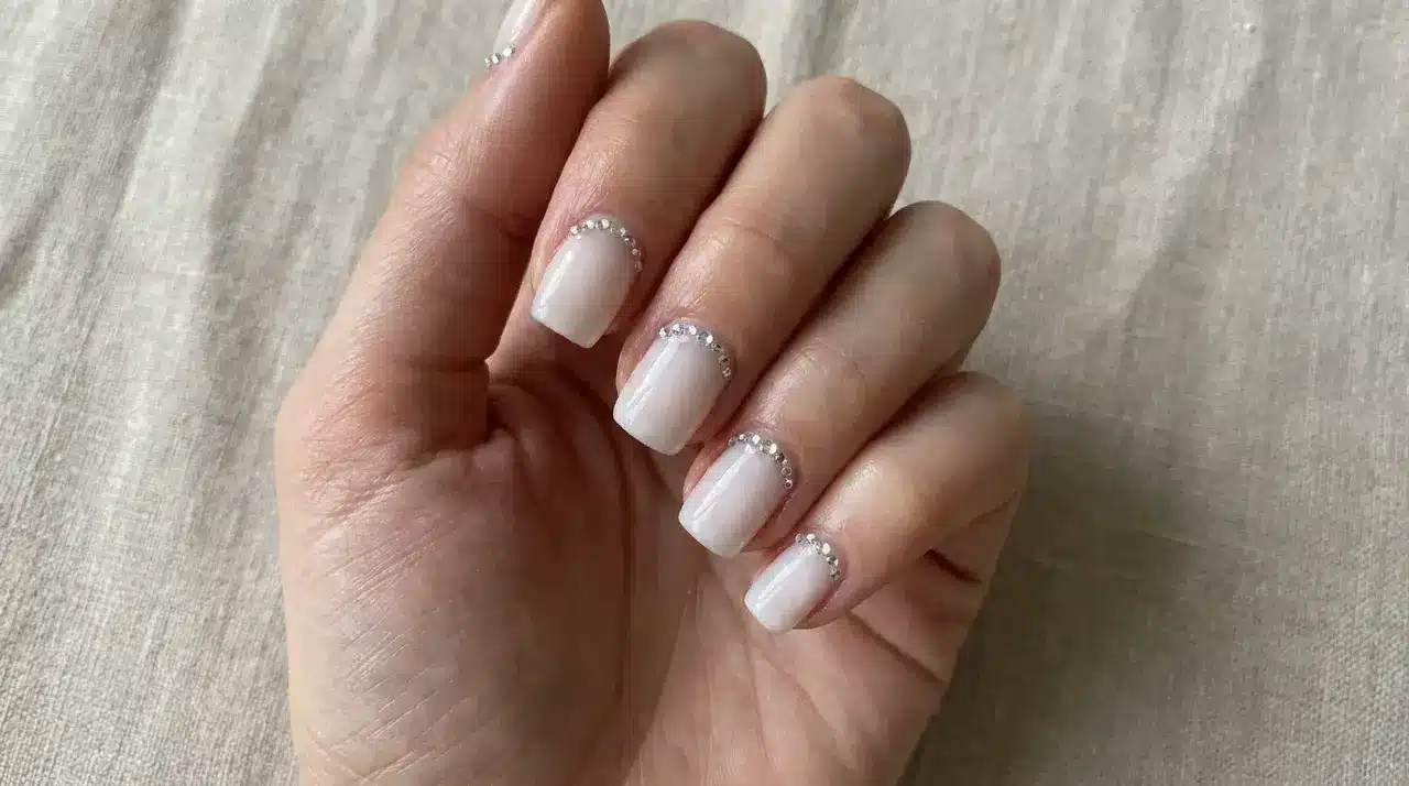 Short square milky white nails with delicate rhinestone cuticle accents on a hand resting over textured fabric background