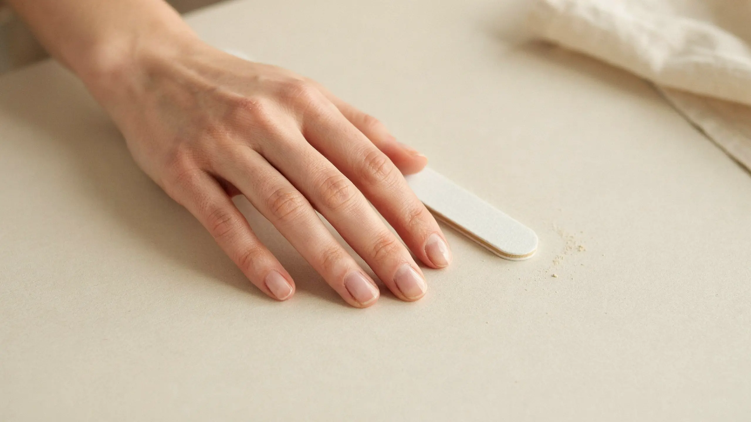 Hand resting beside a white nail file on a beige surface with short natural nails prepared for a basic home manicure set