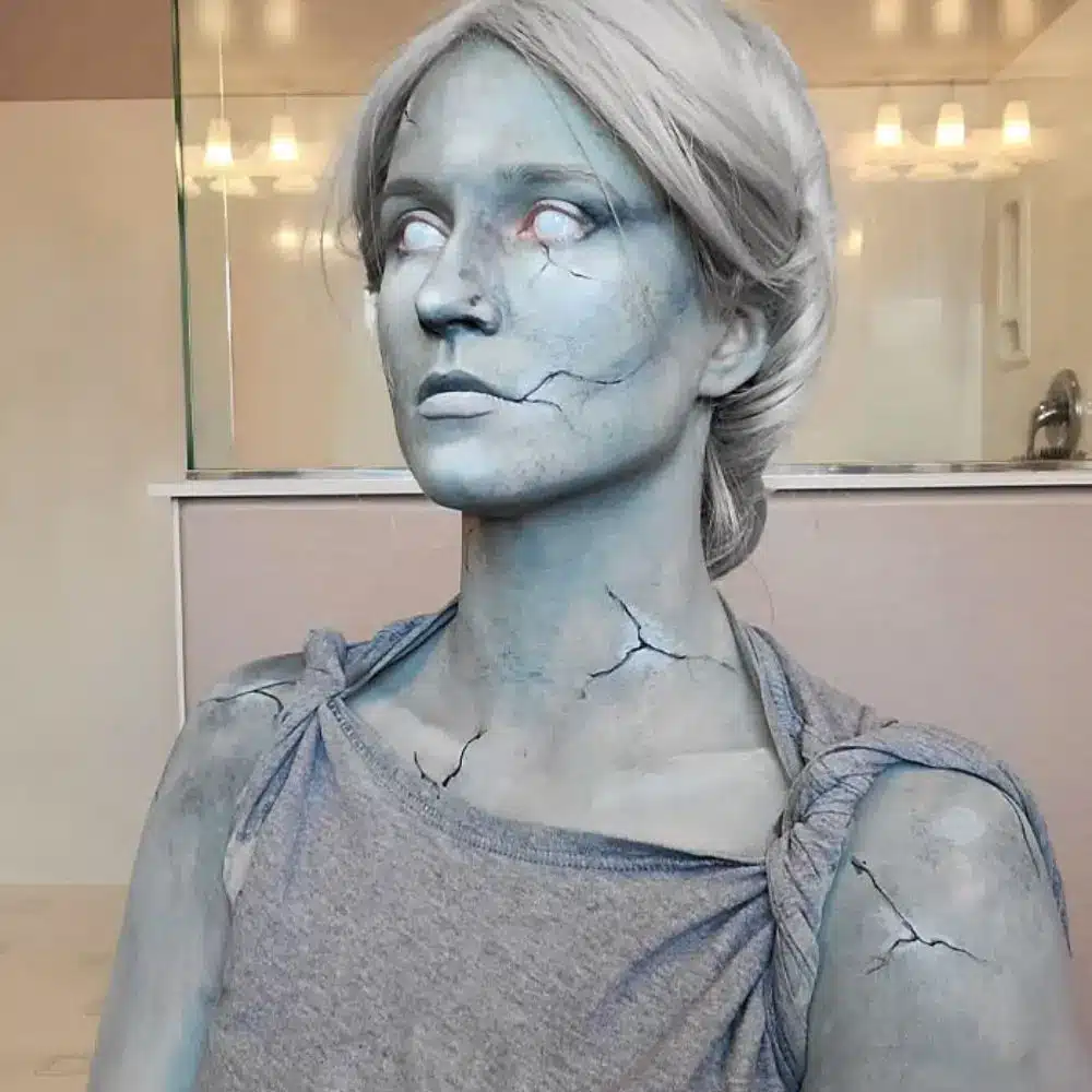Stone face makeup with even grey skin, rough stippled texture, and fine black and white cracks across face and neck