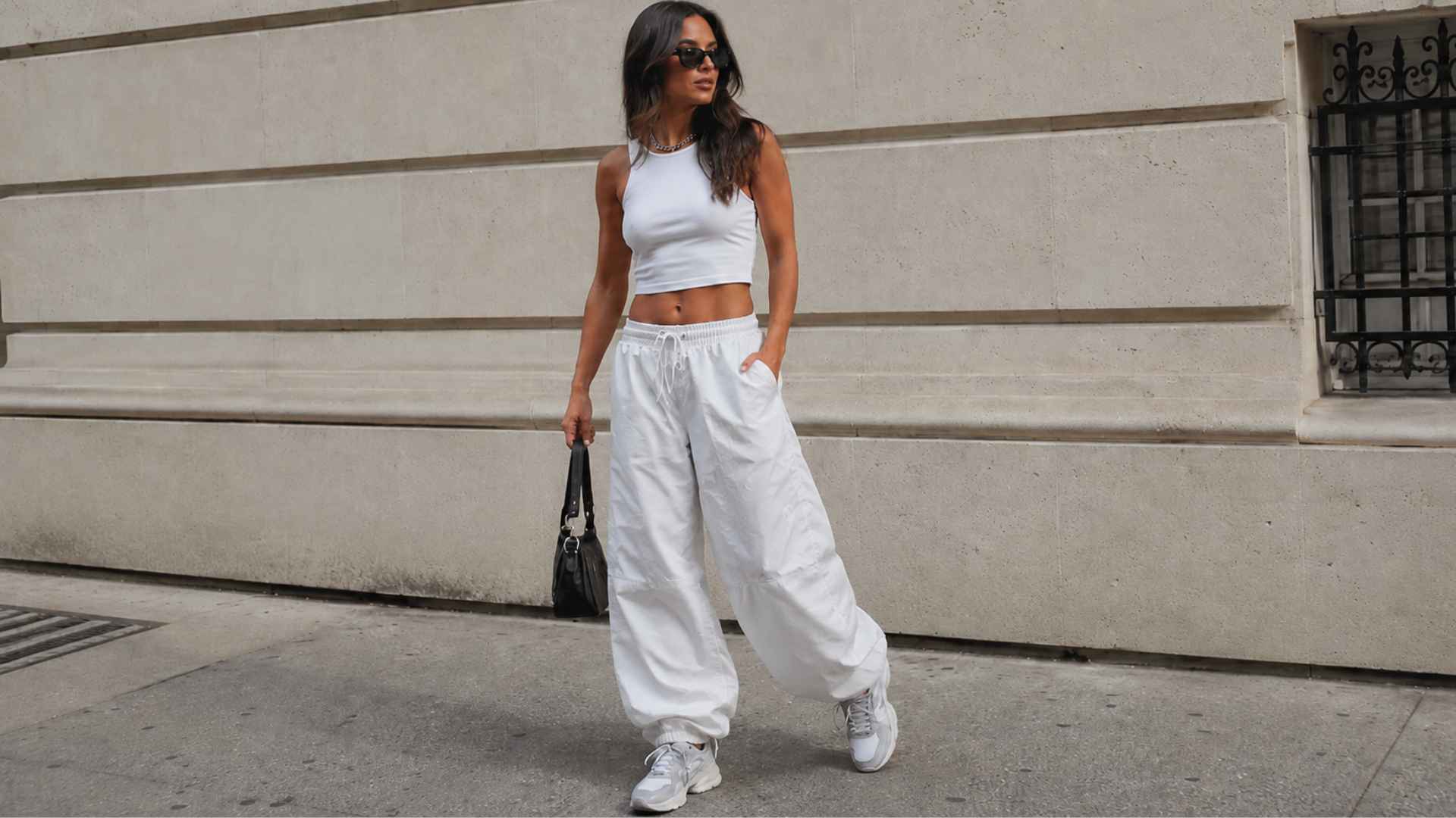 Woman wearing white parachute pants with a fitted crop top, sneakers, sunglasses, and a black bag on a busy city street.