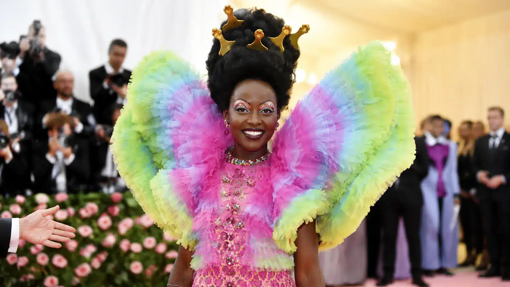 Camp fashion showcased through a colorful ruffled couture look with oversized pastel wings and dramatic crown styling on carpet