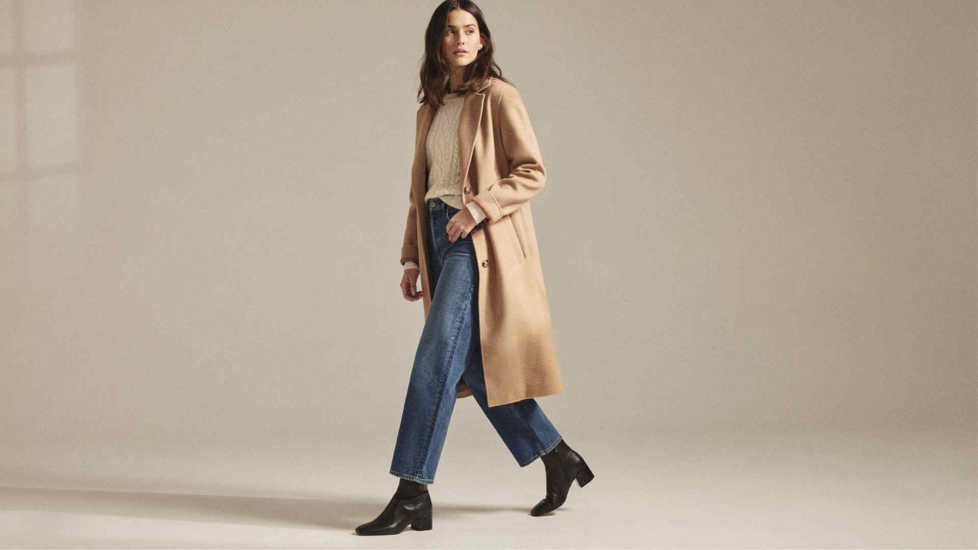 Woman styling wide-leg jeans with a cream sweater, camel wool coat, and ankle boots on a winter city street